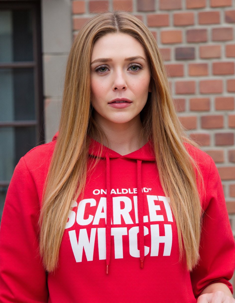 The image is a high-resolution photograph featuring a young woman with long, straight, blonde hair cascading past her shoulders. She has a fair complexion and is wearing a red  "Scarlet Witch" hoodie with a large, white "Scarlet Witch" logo on the front. Her expression is neutral, with slightly parted lips and a direct gaze into the camera. She has well-groomed eyebrows, long, dark eyelashes, and subtle makeup, including a touch of mascara and light lipstick.The background consists of a brick wall with a large, square, glass window partially visible on the left side. The window has vertical bars and is framed by a stone ledge. The bricks are a mix of light and dark shades, contributing to a rustic, urban feel. The lighting is natural, suggesting the photo was taken outdoors during the daytime. The overall composition focuses on the subject, with the background providing a textured, urban context. The photograph captures a blend of casual and stylish elements, emphasizing the woman's appearance and the setting,,Elizabeth Olsen,<lora:Elizabeth Olsen_Flux_V1:0.92>