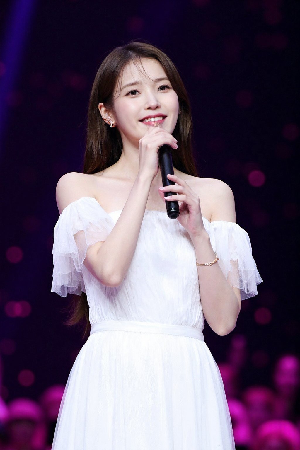 (medium full shot), beautiful young korean girl performing on stage, bathed in bright spotlight. She is wearing a white, off-shoulder dress with flowing, layered fabric, giving her an ethereal and elegant appearance. Her long hair cascades over her shoulders, and she holds a microphone close to her mouth, seemingly in the midst of a song. The background is dark with a bokeh effect of small pink and purple lights, suggesting a large audience or a concert setting. , <lora:flux_realism_lora:1>,  <lora:makinaflux_iu_v1.0:1>