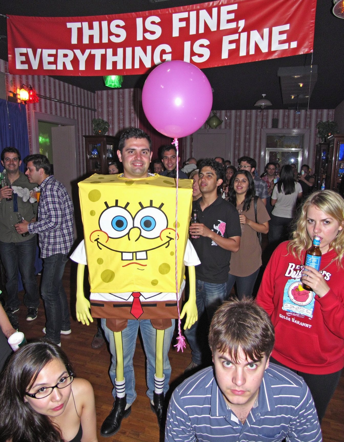 In a wide shot of a night club, a man dressed as the grumpy character resembling SpongeBob is holding a balloon. Above him, a banner reads: “THIS IS FINE. EVERYTHING IS FINE.” People drinking happily while he stands there, clearly not having it <lora:amateurphoto-v6-forcu:0.8>