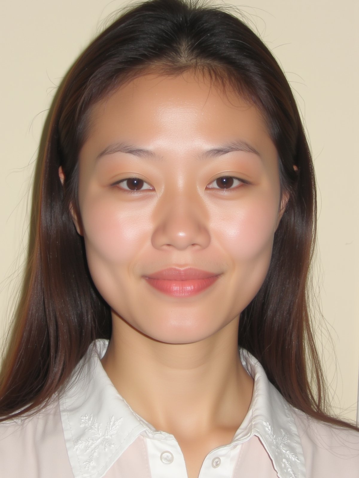 A close-up shot of a young Asian woman with long dark brown hair and brown eyes. She is wearing a white collared button down shirt with white embroidery on the collar. Her lips are a vibrant shade of pink. Her eyebrows are a light brown and her eyes are a darker shade of brown. She has a slight smile on her face. The backdrop is a light beige wall.