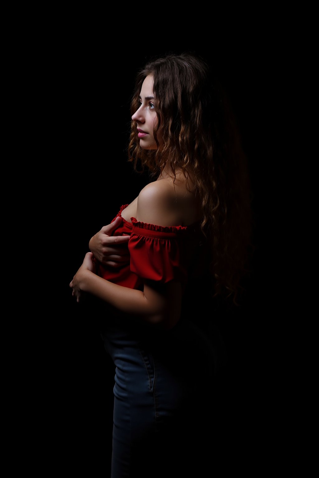 The image is a portrait of a young woman with long, curly hair. She is wearing a red off-shoulder top and blue jeans. Her arms are crossed in front of her chest and she is looking off to the side with a thoughtful expression on her face. The background is completely black, making the woman the focal point of the image. The lighting is soft and dramatic, creating a moody atmosphere