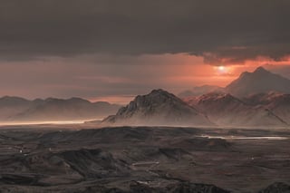 The image depicts a dramatic and industrial landscape. A thick layer of gray clouds dominates the sky, illuminated in a deep red hue from above. Amidst the clouds, a small golden sun peeks out, adding a touch of warmth to the otherwise gloomy atmosphere. In the distance, on the horizon, a range of gray mountains stretches across the landscape. Among these mountains, power plants and industrial buildings can be seen, their structures blending into the rugged terrain. Closer to the foreground, the mountains take on a darker tone, gradually transitioning to black mountains that loom in the distance. These black mountains are also dotted with power plants and industrial buildings, casting long shadows in the fading light.