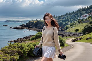 The photo shows a young woman standing on a rocky beach.  The composition of the image is casual and candid.  The subject wore a beige sweater and a white skirt, showing off a sexy and charming smile.  Embodying a feeling of happiness and freedom.  She is carrying a brown cross-body bag.  The background includes a tranquil coastal landscape, a rocky shoreline, calm ocean waves, and distant forested hills under a partially cloudy sky.  There is a walking path with stone walls on the right side of the image, indicating a coastal path or promenade.  The overall mood is relaxed and cheerful.