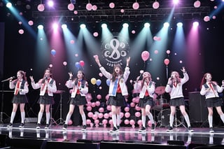 A student in a school uniform stood on the stage holding a guitar and singing with a microphone. The stage was surrounded by five students, three of whom were holding balloons and two of whom- were holding banners. The atmosphere was very lively.

